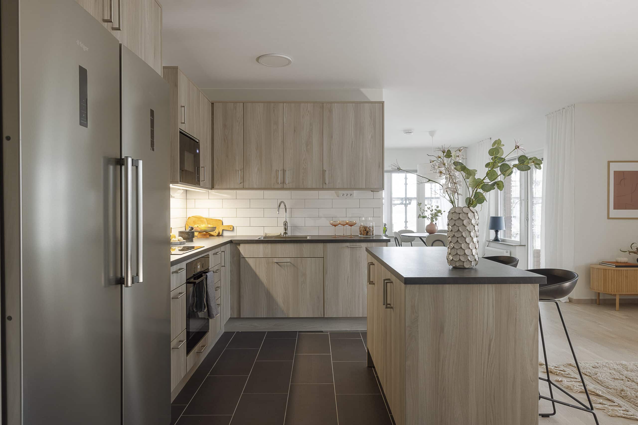Modern kitchen with light wood cabinets, stainless steel fridge, and a dark tile floor.