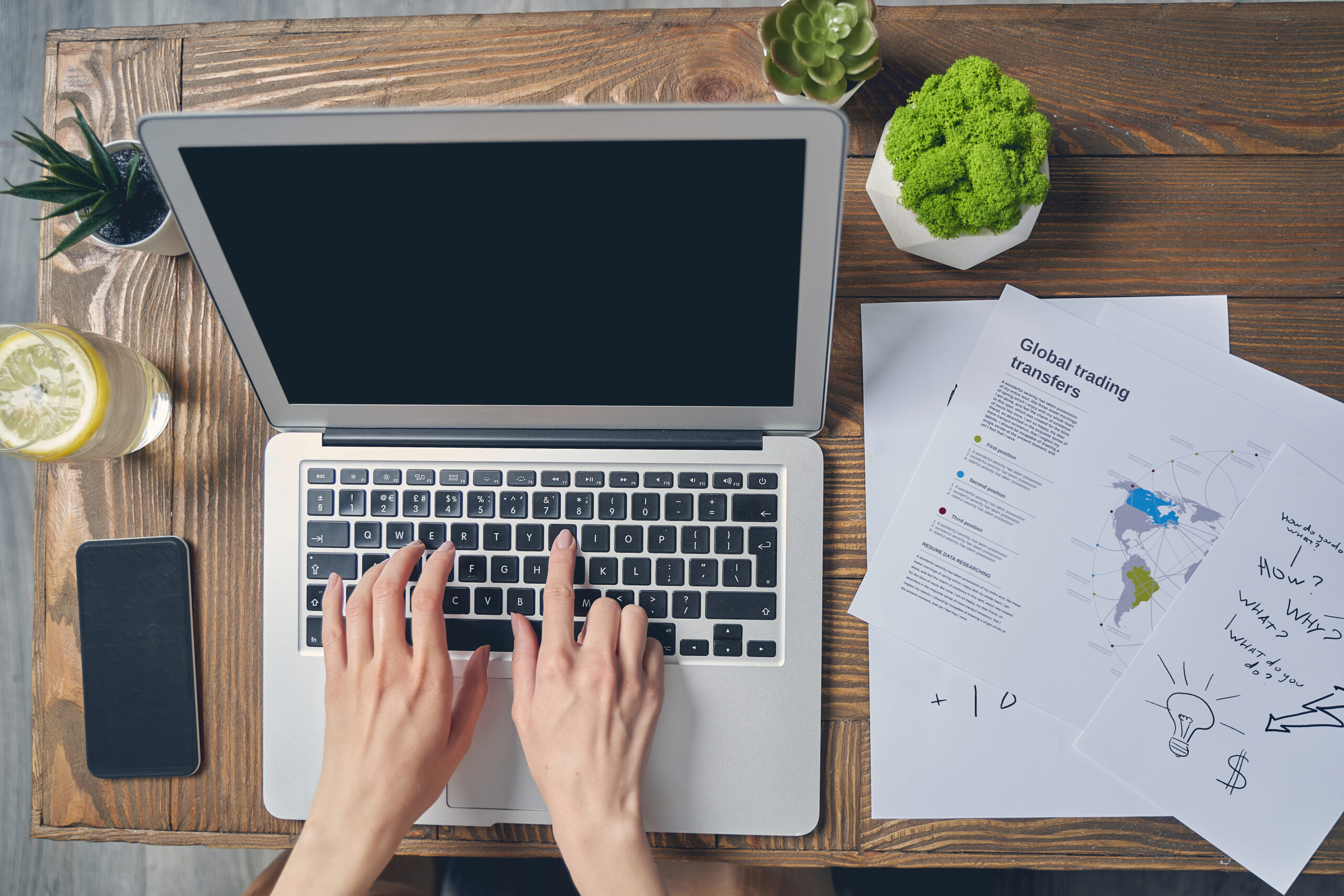 Beautiful female hands typing on laptop keyboard Top down view of a person typing on a laptop at a wooden desk with trading documents a smartphone a lemon drink and potted plants nearby